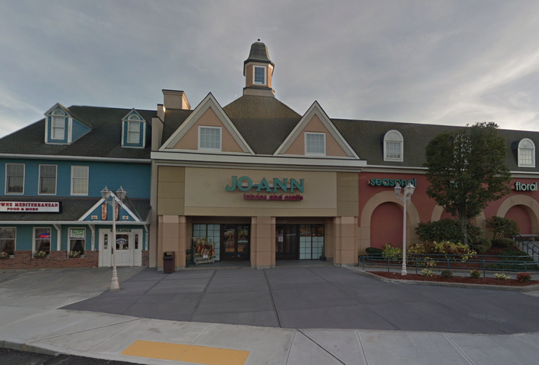 A Joann store sits within a row of strip mall stores behind a paved walkway.