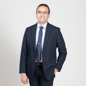 A man wearing glasses stands with one hand in his pocket while wearing a dark blue suit.