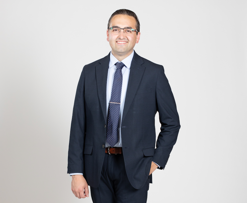 A man wearing glasses stands with one hand in his pocket while wearing a dark blue suit.