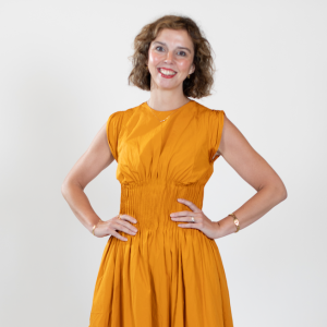 A woman with short blonde hair wears a mustard-colored dress, standing with her hands on her hips.