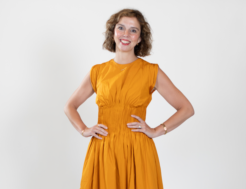 A woman with short blonde hair wears a mustard-colored dress, standing with her hands on her hips.