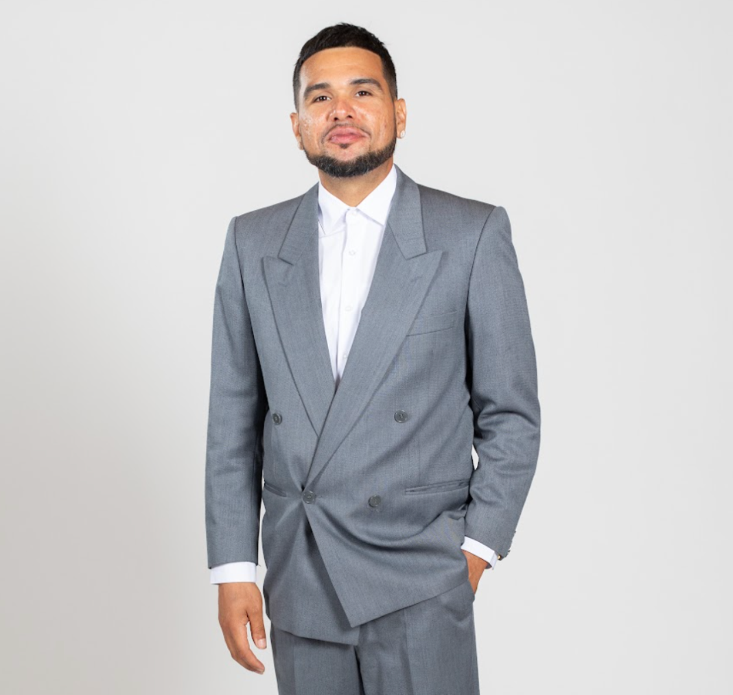 A man wears a grey suit and stands with his hand in his pocket.