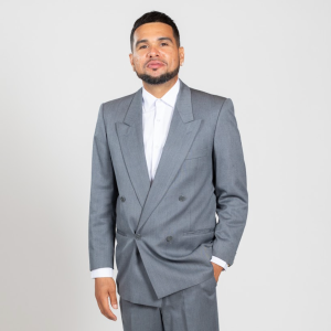 A man wears a grey suit and stands with his hand in his pocket.