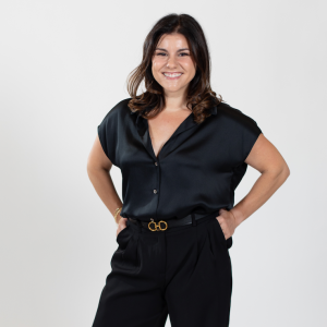 A woman with shoulder length brown hair stands with her hands in her pant pockets wearing a black top and pants.