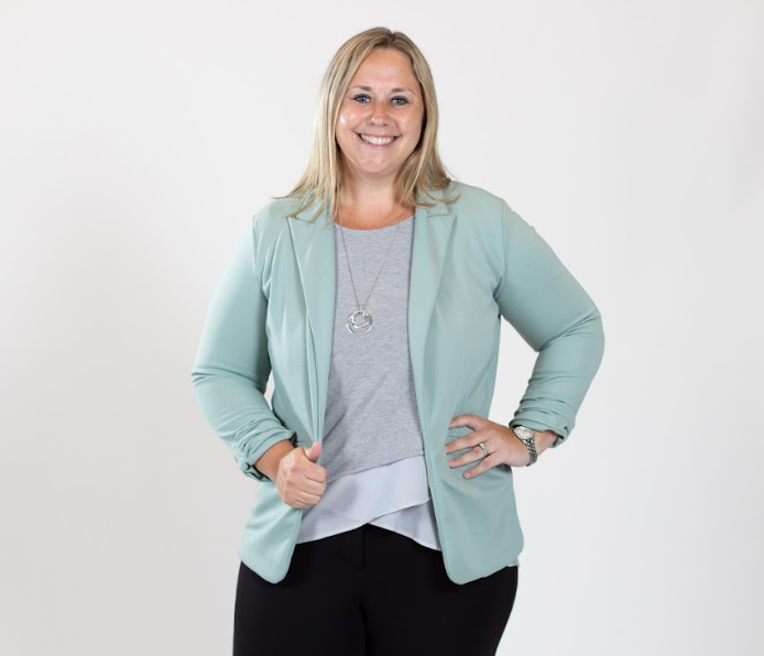 A woman with blonde hair wears a grey bouse and a pale blue blazer.
