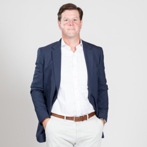 A man stands wearing a white shirt and pants with a blue blazer and his hands tucked into his pockets.