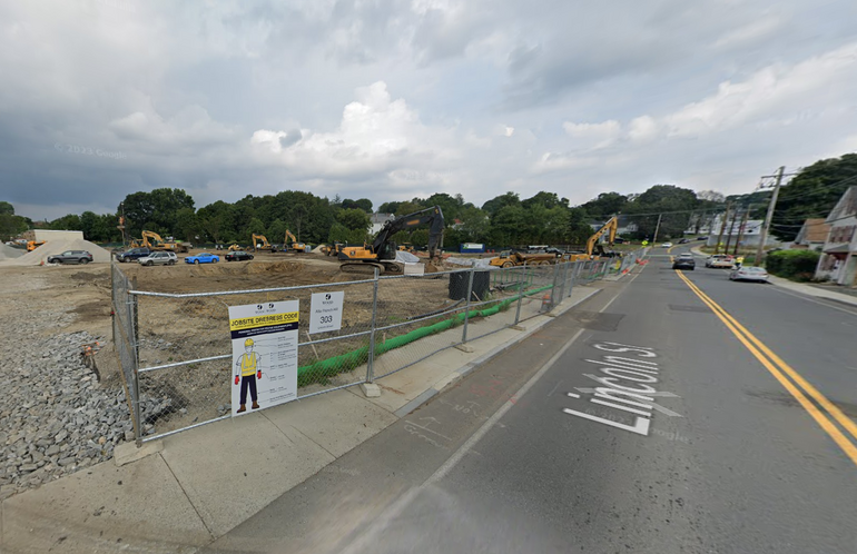 A fence with dirt and construction materials behind it. The fence sits on the sidewalk of a two lane road.