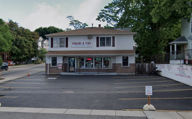 A two story building, with a retail shop on bottom and apartment windows on the top floor. An empty parking lot sits in front.