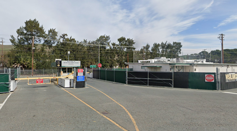 A gate at the front of a military base, with a green fence with barbed wire to the right of the gate.