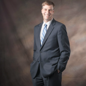 A man with brown hair standing in a dark suit, striped button down, and blue diagonally striped tie.