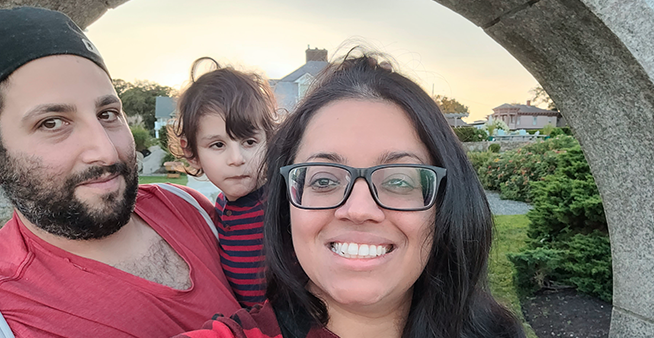 A man in a black baseball cap and red t-shirt holds a boy in a red and black striped sweater next to a woman with black glasses and long dark hair smiling for the camera.