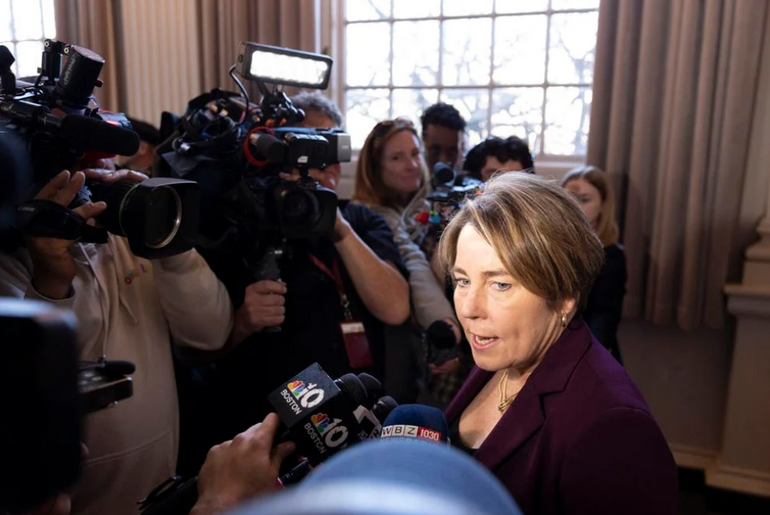 A woman with short brown and blonde hair wears a purple blazer while speaking to reporters.