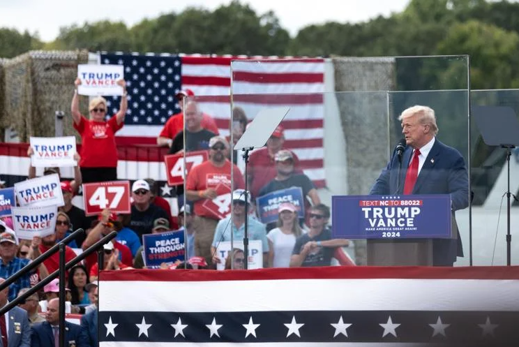 Man behind bullet-proof glass speaks on a stage at a political rally