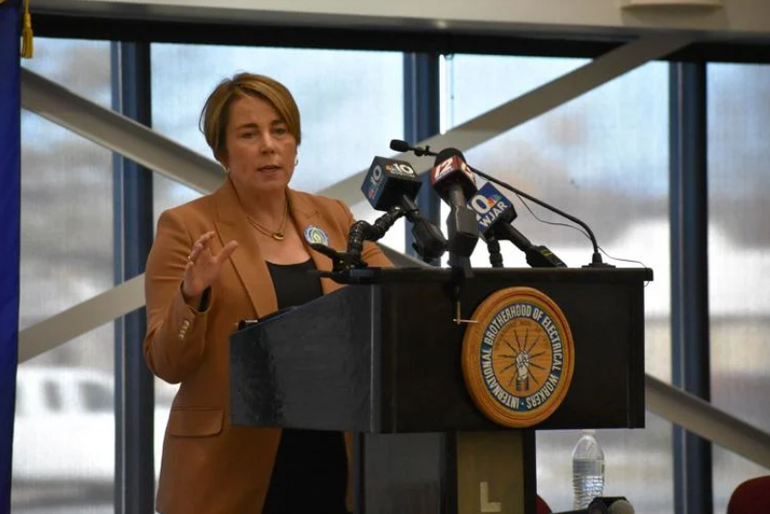 A woman with short blonde hair wears a tan suite jacket and black top while speaking behind a podium with several microphones on it.