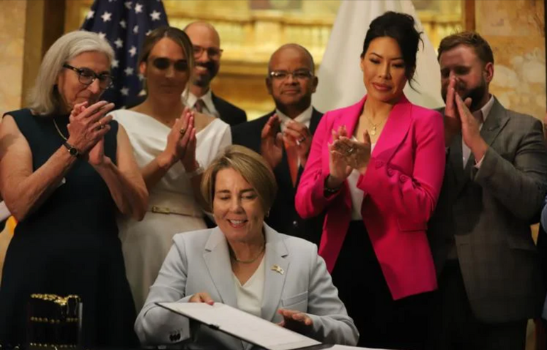 A woman sits at a table holding a piece of legislation as people behind her clap.