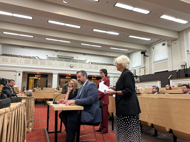 Two people sit at a table with a microphone at a hearing in an auditorium