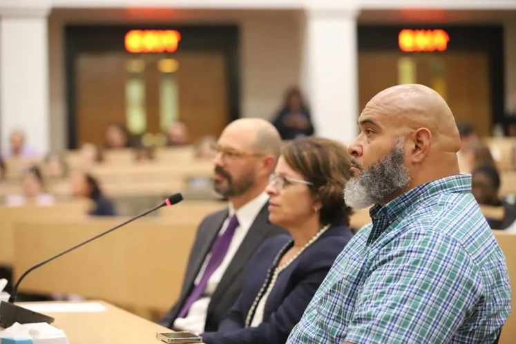 Three people in business attire sit at a table with a microphone during a hearing