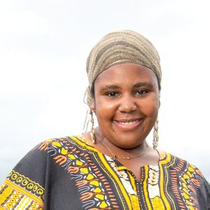 A woman wears a green patterned headwrap and a black, yellow, orange, and white patterned top.