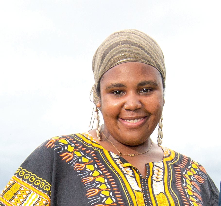 A woman wears a green patterned headwrap and a black, yellow, orange, and white patterned top.