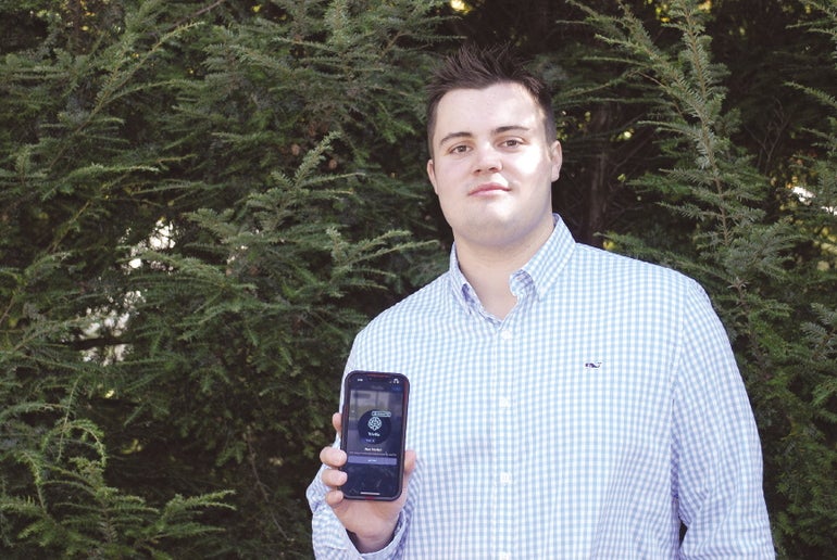 A man in a dress shirts stands in front of foliage holding up a phone to the camera