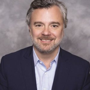 A man with grey hair and beard wears a dark blue suit jacket and light white button down.
