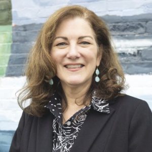 A woman with brown hair wears a black suit jacket and black and white patterned top.