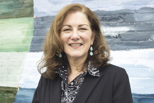 A woman with brown hair wears a black suit jacket and black and white patterned top.