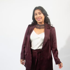 A woman with long black hair wears a black beaded necklace, a white satin top, and maroon satin suit jacket and pants.