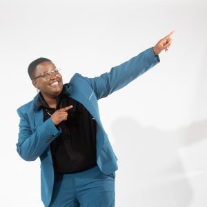 A woman with glasses in a blue suit points upwards and to the right with a smile on their face