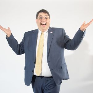 A man with glasses shrugs while smiling in a suit
