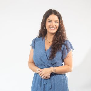 A woman with long curly brown hair wears a blue dress, hoop earrings, and a silver heart necklace.