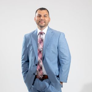 A man wears a light blue suit jacket and pants, a brown belt, a white button down, and a pink, grey, and white patterned tie with his hands in his pockets.