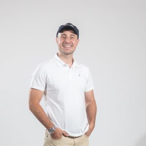 A man in a baseball hat and white polo with his hands in his pockets