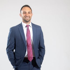 A man wears a blue suit jacket and pants, a white button down, and a purple and red paisley tie.