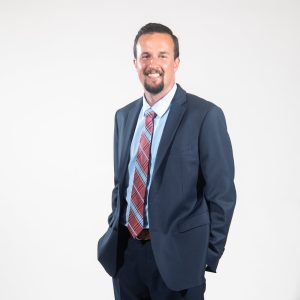 A man with short brown hair and a goatee wears a dark blue suit jacket and pants, a light blue button down, and a red and light blue plaid tie with his hands in his pockets.