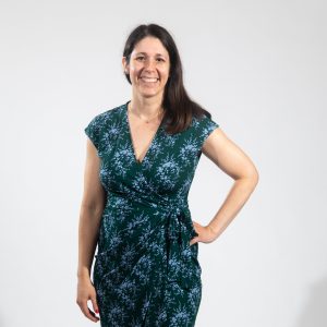 A woman with long brown hair wears a green and blue patterned dress, red nail polish, and one hand on her hip.