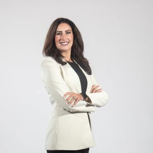 A woman with long brown hair wears a black top and white blazer, standing with her arms folded.