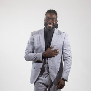 A man wears a grey suit, vest, and pants, and a black button down with one hand on his chest.
