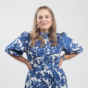 A woman with curled blonde hair wears a blue and white floral dress with elbow length sleeves.