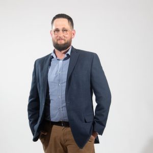 A man wears a dark blue suit jacket, light blue button down, and light brown pants with his hands in his pockets.