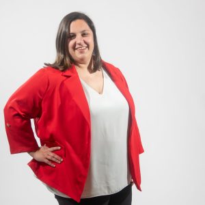 A woman with shoulder length brown hair wears a white top and red blazer with her hand on her hip.