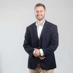 A man wears a black suit jacket, a white button down, and tan pants.