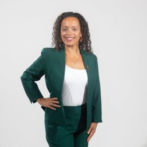 A woman with long curly hair wears a dark green suit jacket and pants and a white top.