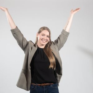 A woman in a grey suit jacket with her hands up in celebration