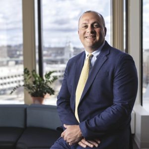 A man sits on a couch with his hands together wearing a blue suit in front of large office windows.