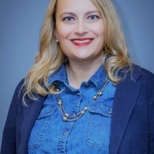 A woman with slightly curled blonde hair wears red lipstick, a blue denim top, a dark blue blazer, and a silver necklace.
