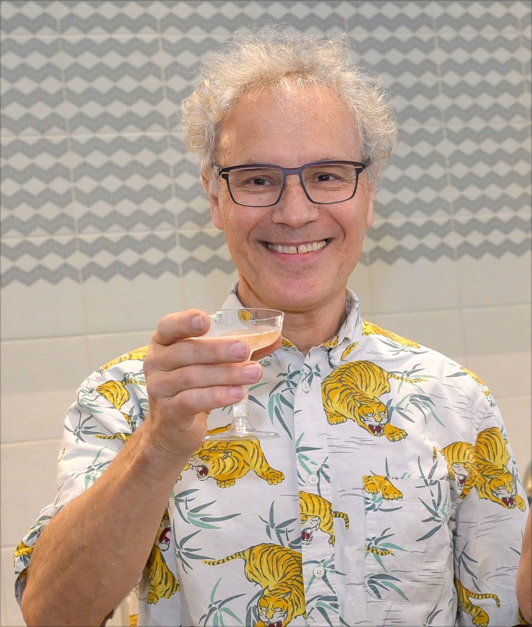 Man in dress shirt with tigers and leafs on it smiles while holding  a martini glass