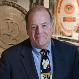 A man wears a dark blue blazer, light blue button down, and a Mickey Mouse tie.