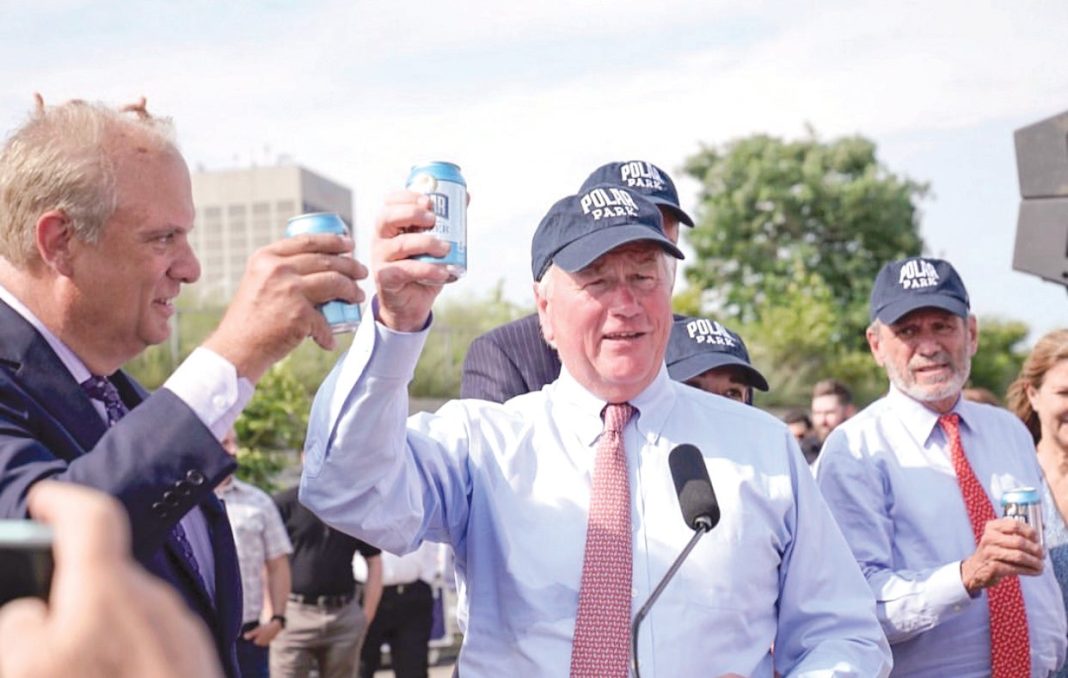 Men in light blue shirts, red ties, and dark blue baseball caps hold up blue cans of seltzer outside.