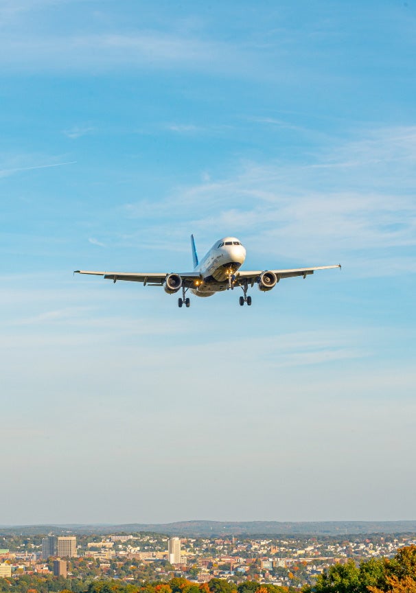 A white plane flies in a light blue sky with slight cloud coverage over a city landscape with trees and hills